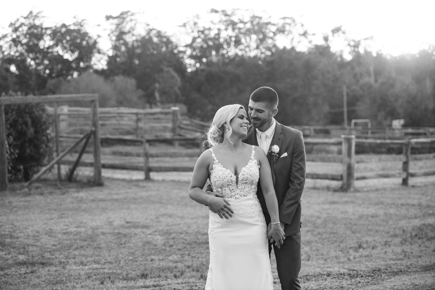 A couple in wedding attire walks together in a rustic outdoor setting with wooden fences at sunset.