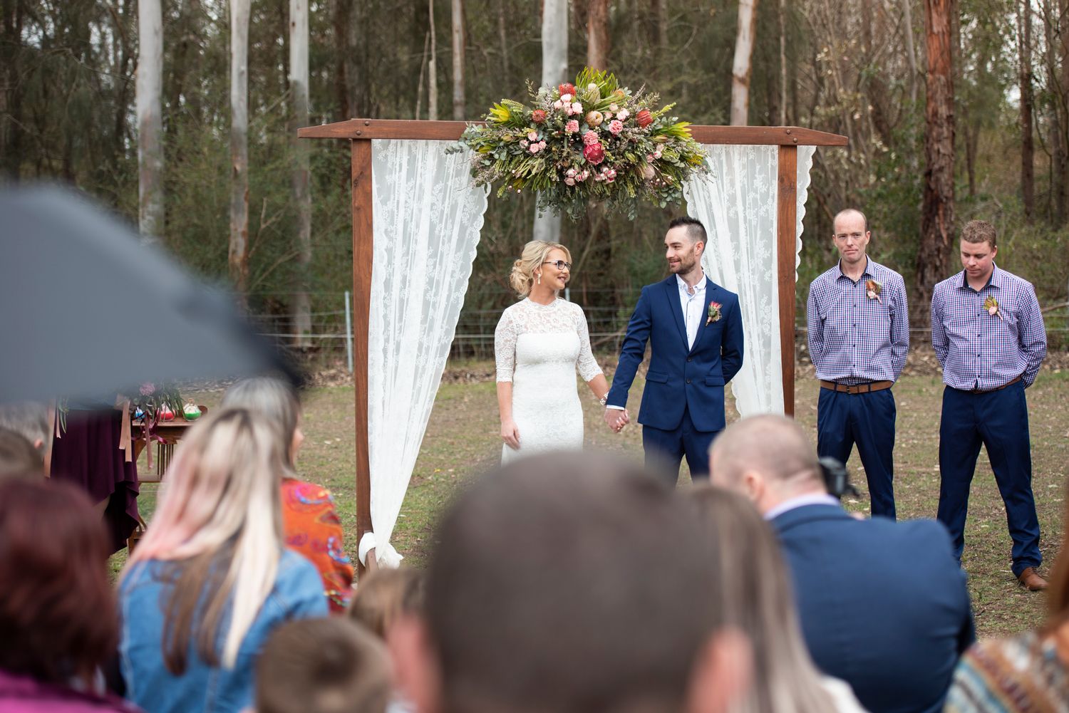 An outdoor wedding ceremony in a country setting with a decorated arch and guests seated on wooden benches.