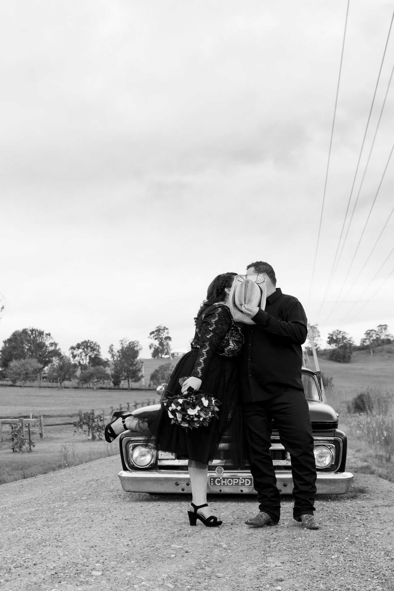 A romantic vintage black and white photo series showing a couple sitting on a classic car in a rural setting.