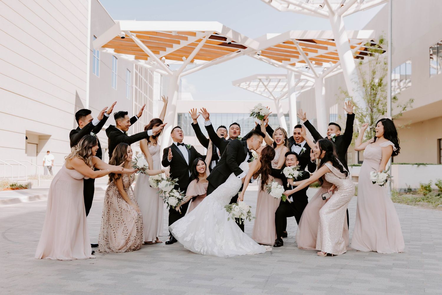 Bridal party group portrait celebrating and cheering for the bride and groom as they share a kiss in Oklahoma City's Scissortail Park.