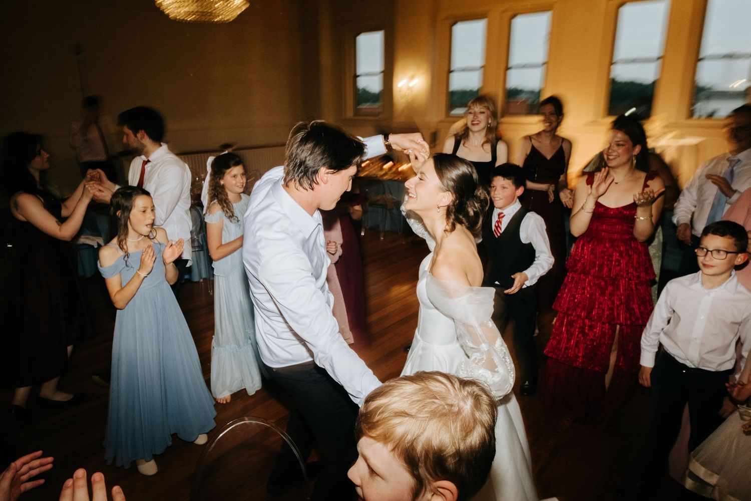 Wedding guests dance and celebrate together on a wooden dance floor at an indoor reception venue.