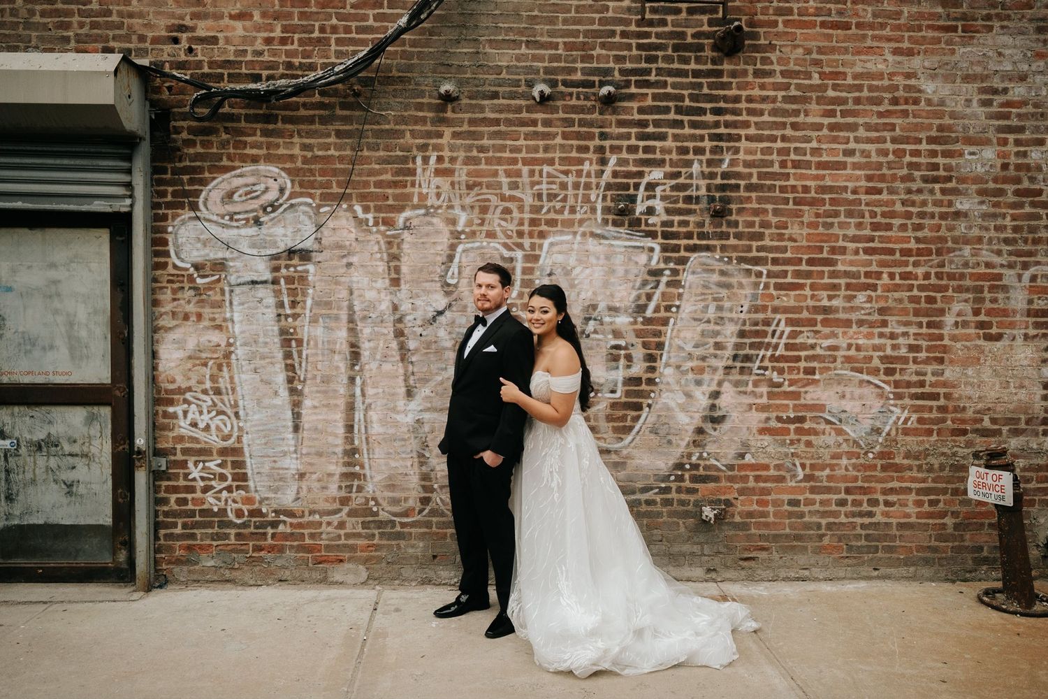 Couple in formal attire poses against rustic brick wall in romantic wedding photoshoot.