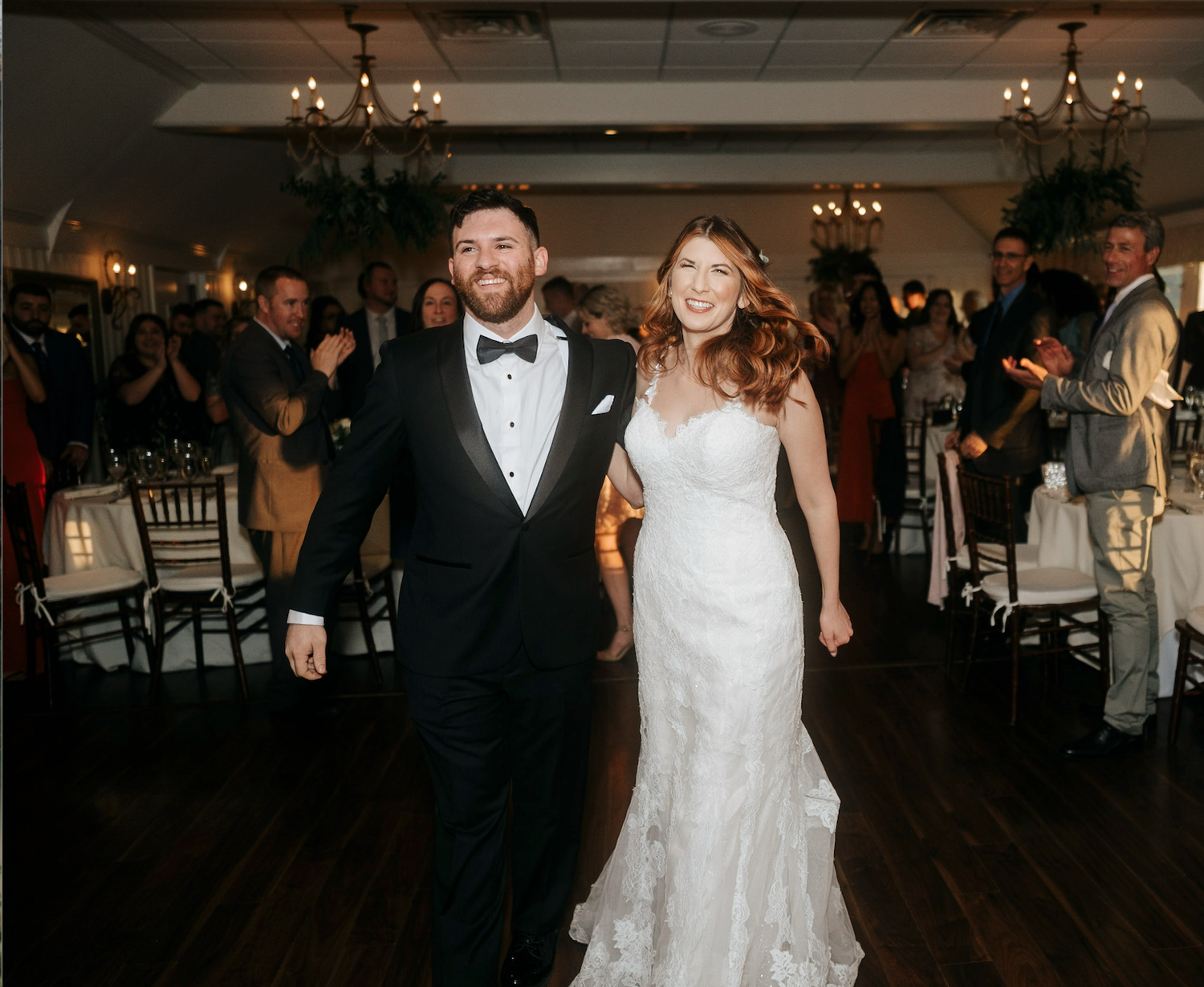 Bride and groom walk hand-in-hand at elegant wedding reception, smiling joyfully as guests applaud in candlelit ballroom.