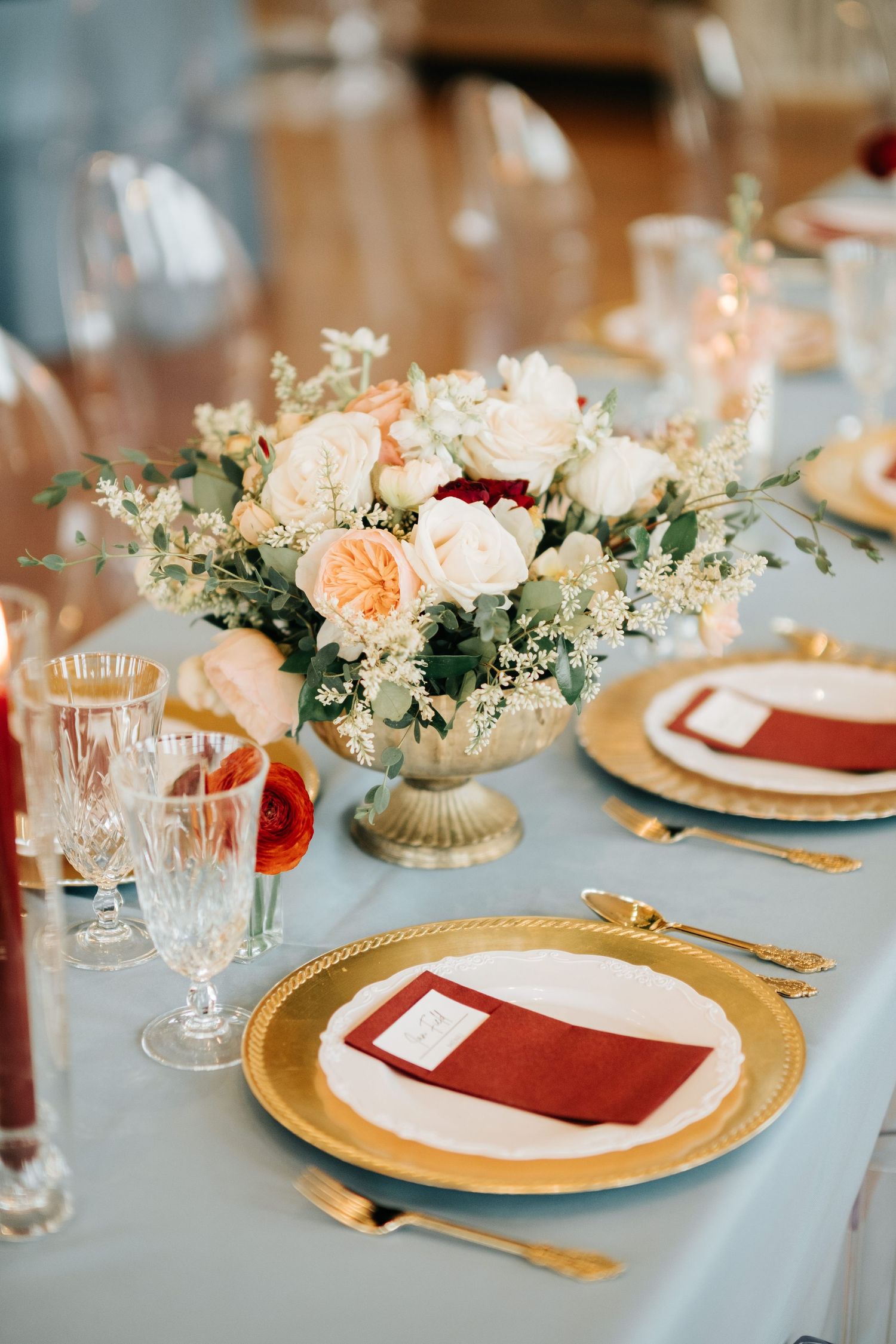 Elegant table settings with gold charger plates, burgundy napkins, and peach floral centerpieces on light blue linens.