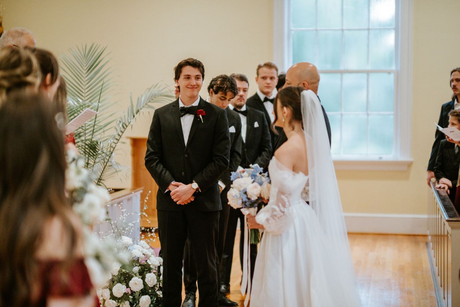 A wedding ceremony takes place in a bright indoor venue with white floral decorations and palm fronds.