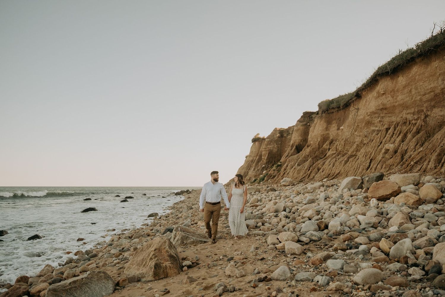 Couple holding hands walking along a rocky beach shoreline with sandy cliffs at golden hour.