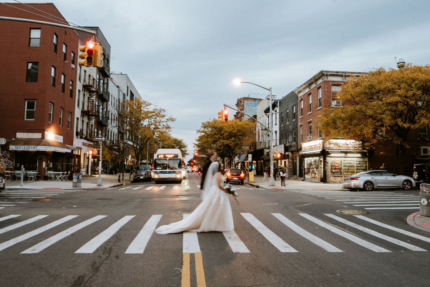 Bride in white gown with long train crosses a busy urban intersection on a cloudy day in New York City.