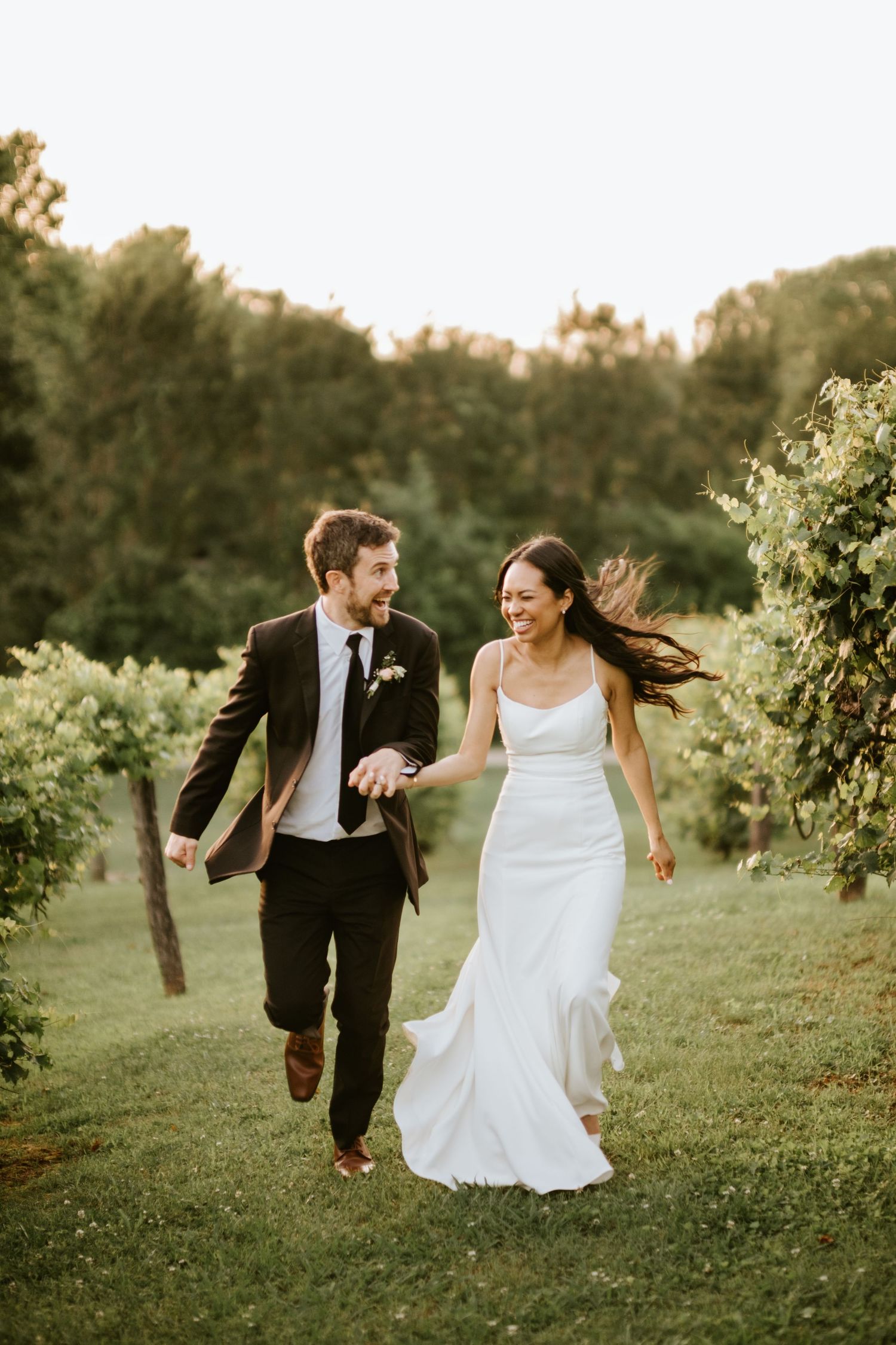 A couple in wedding attire runs joyfully through a vineyard at sunset, creating a romantic and candid moment.