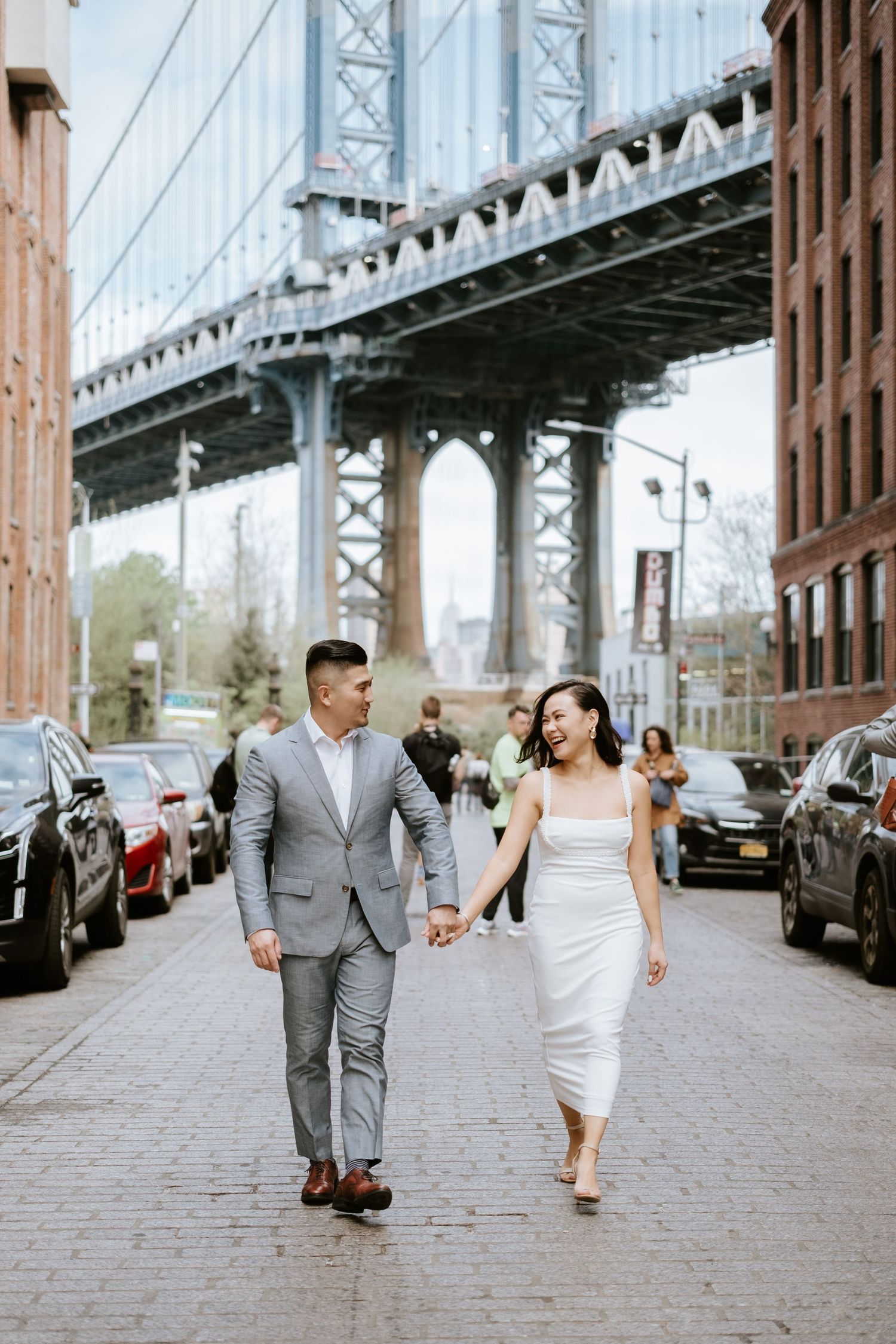 A couple in formal attire walks together on a cobblestone street beneath the Manhattan Bridge in DUMBO, Brooklyn.
