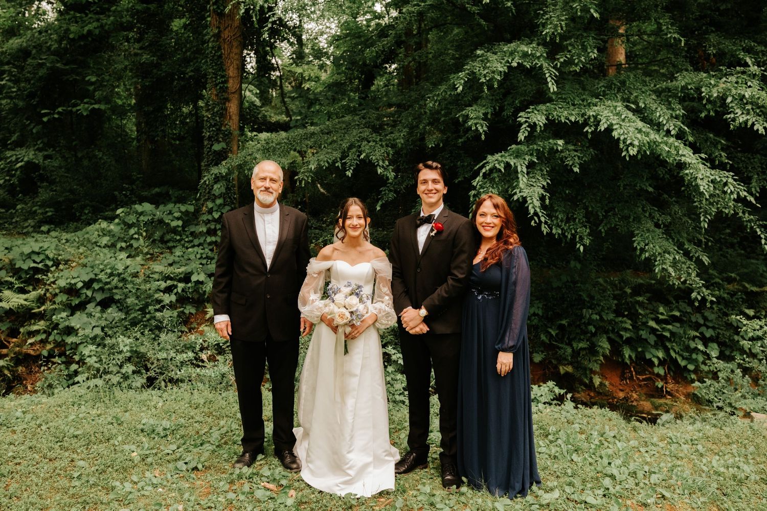 A wedding party stands in a lush forest setting wearing formal attire in shades of navy and white.