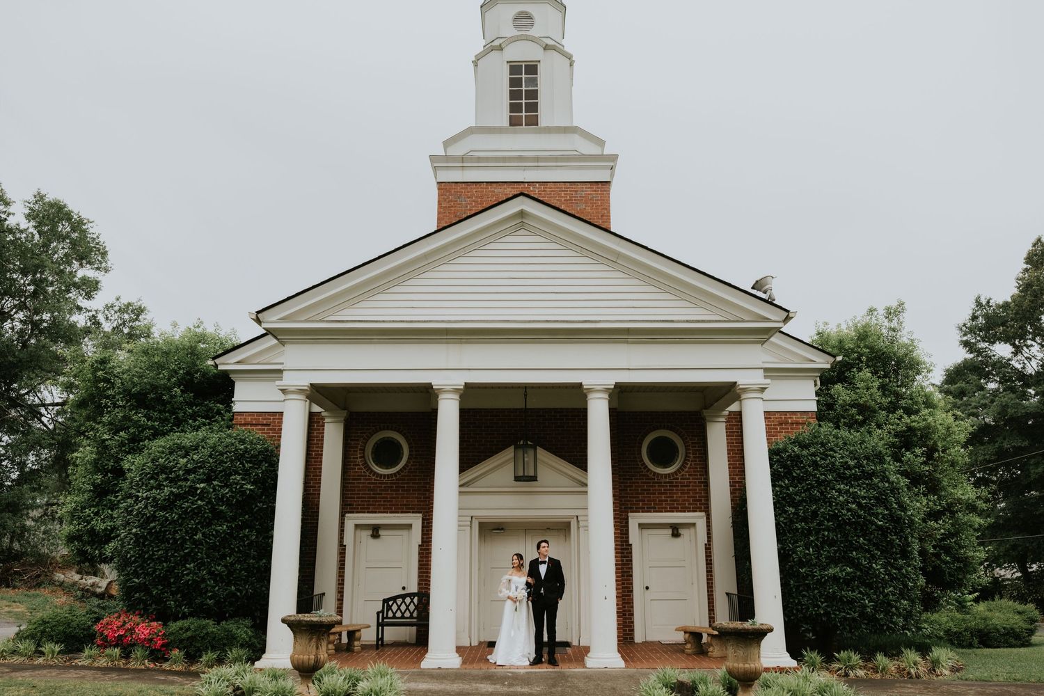 A classic white church with columns and steeple stands elegantly against green trees on a cloudy day.
