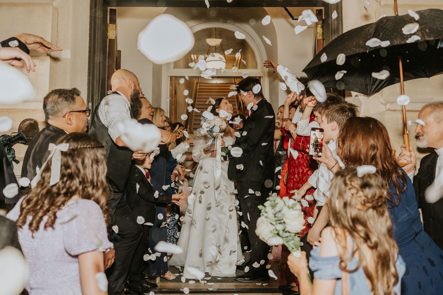 Newlywed couple exits wedding venue through cheering guests tossing white flower petals under umbrellas.