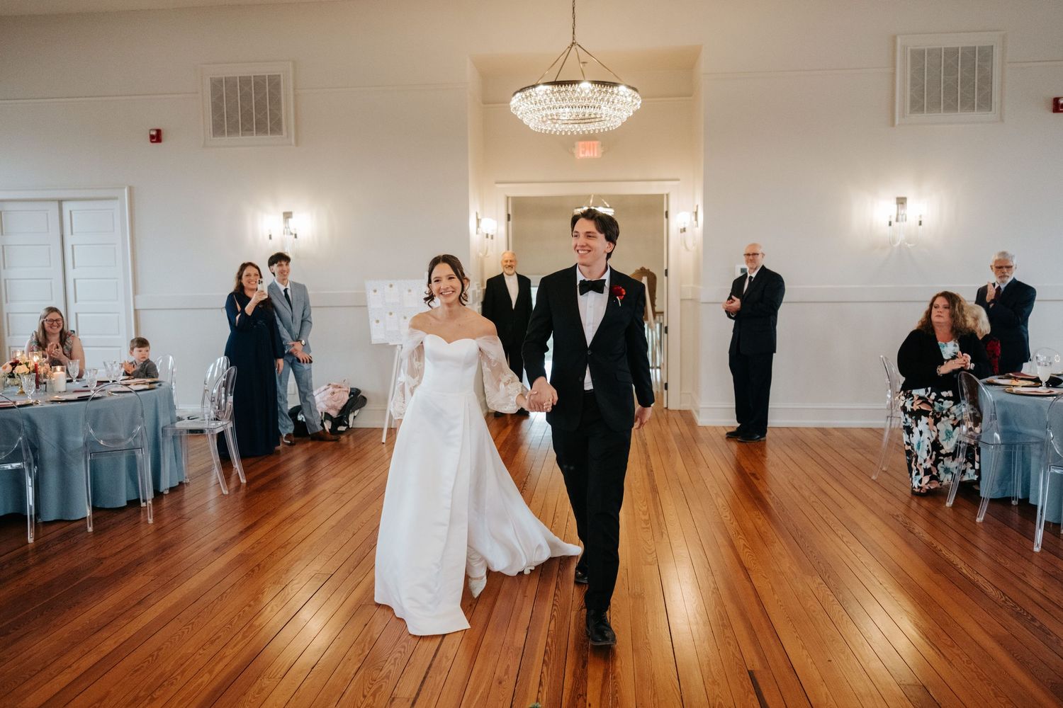 A couple shares their first dance on a polished hardwood floor in an elegant white reception venue with guests watching.
