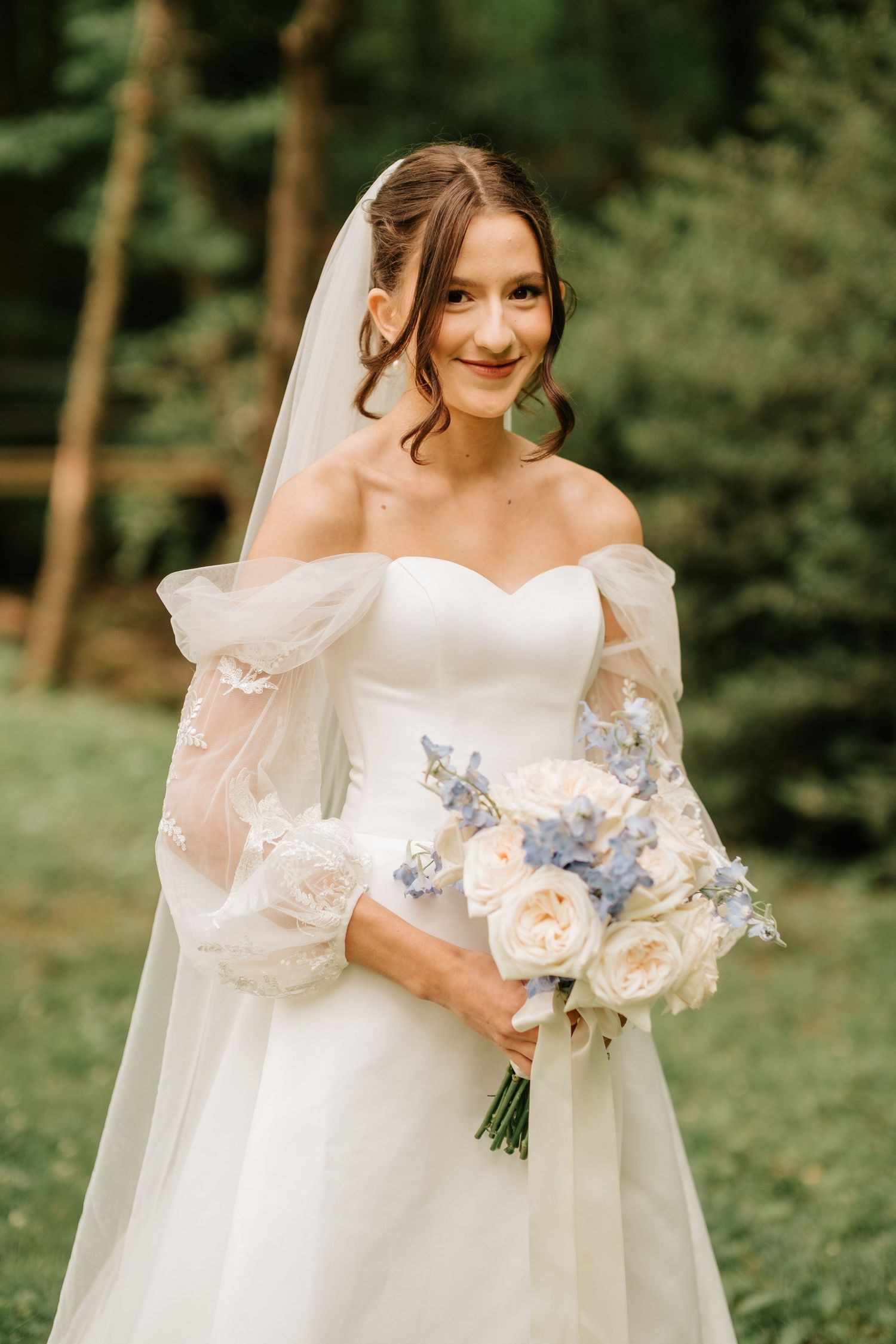 A bride in an off-shoulder white wedding dress holds a romantic bouquet of white and blue flowers in a forest setting.