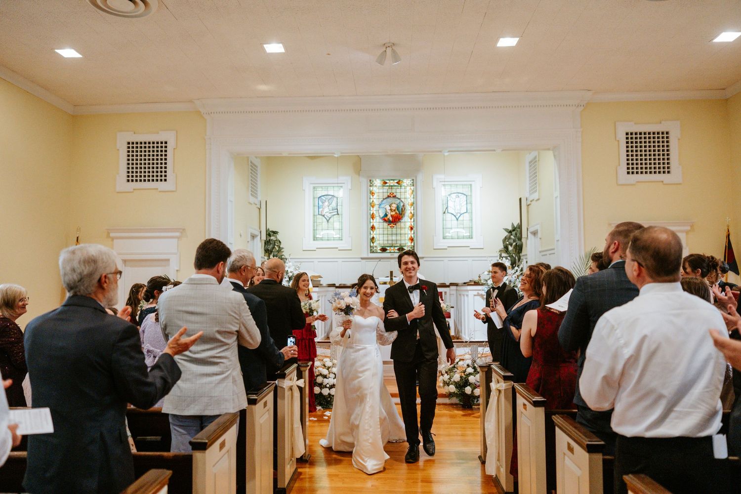 Wedding guests dancing and socializing in an elegant reception hall with white decor and recessed lighting.