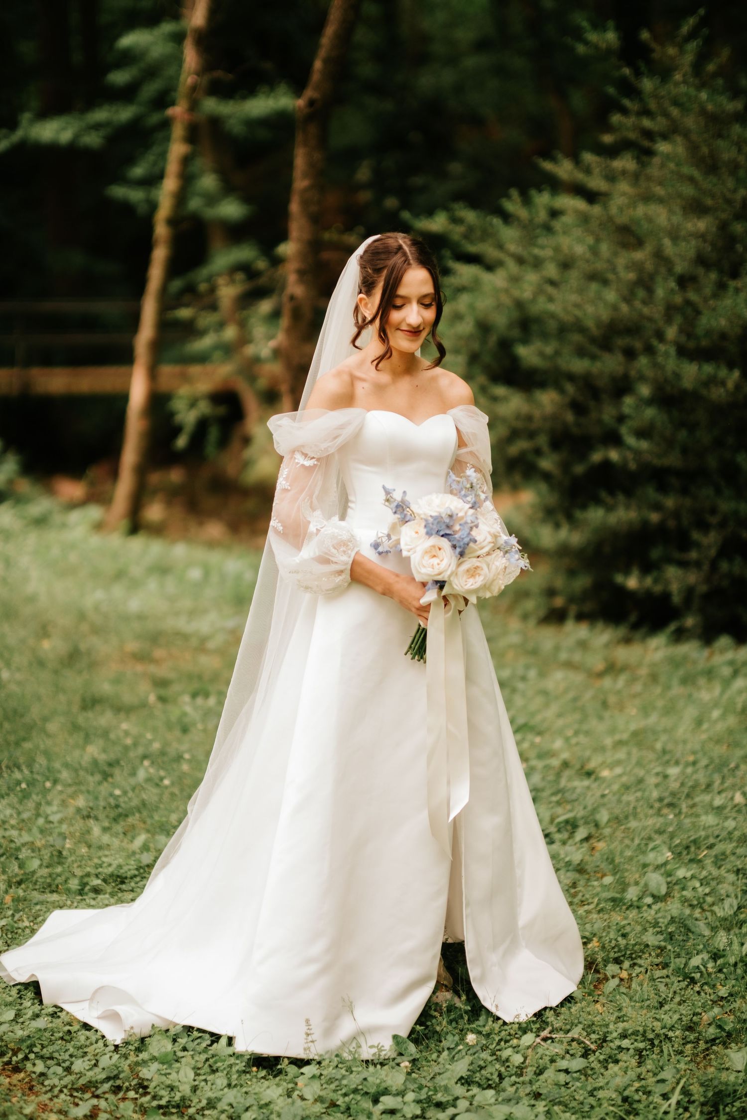 A bride in a flowing white wedding gown stands gracefully in a wooded outdoor setting surrounded by greenery.