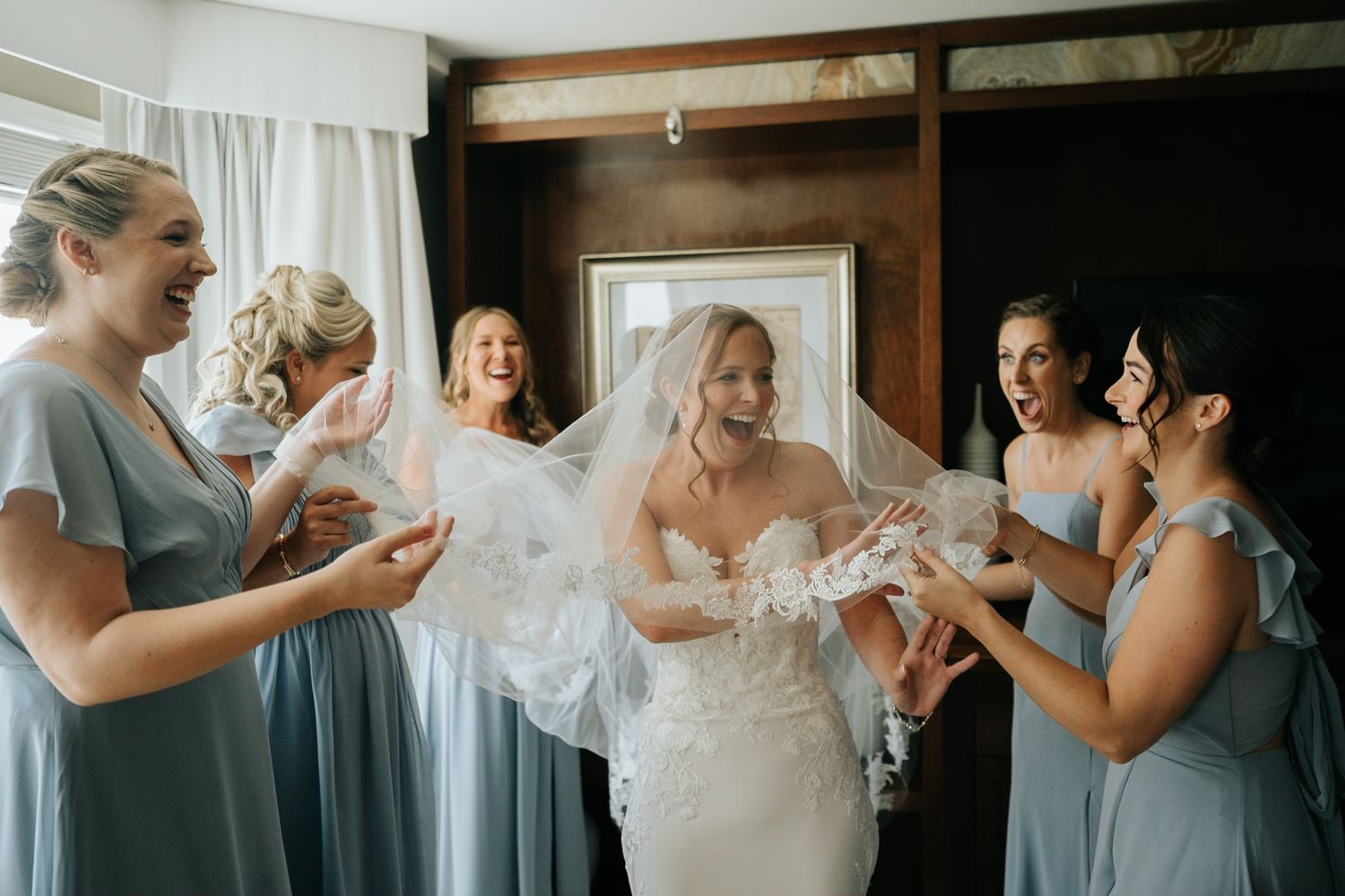 Bride in lace wedding dress and veil laughs with bridesmaids in blue dresses during wedding preparations.