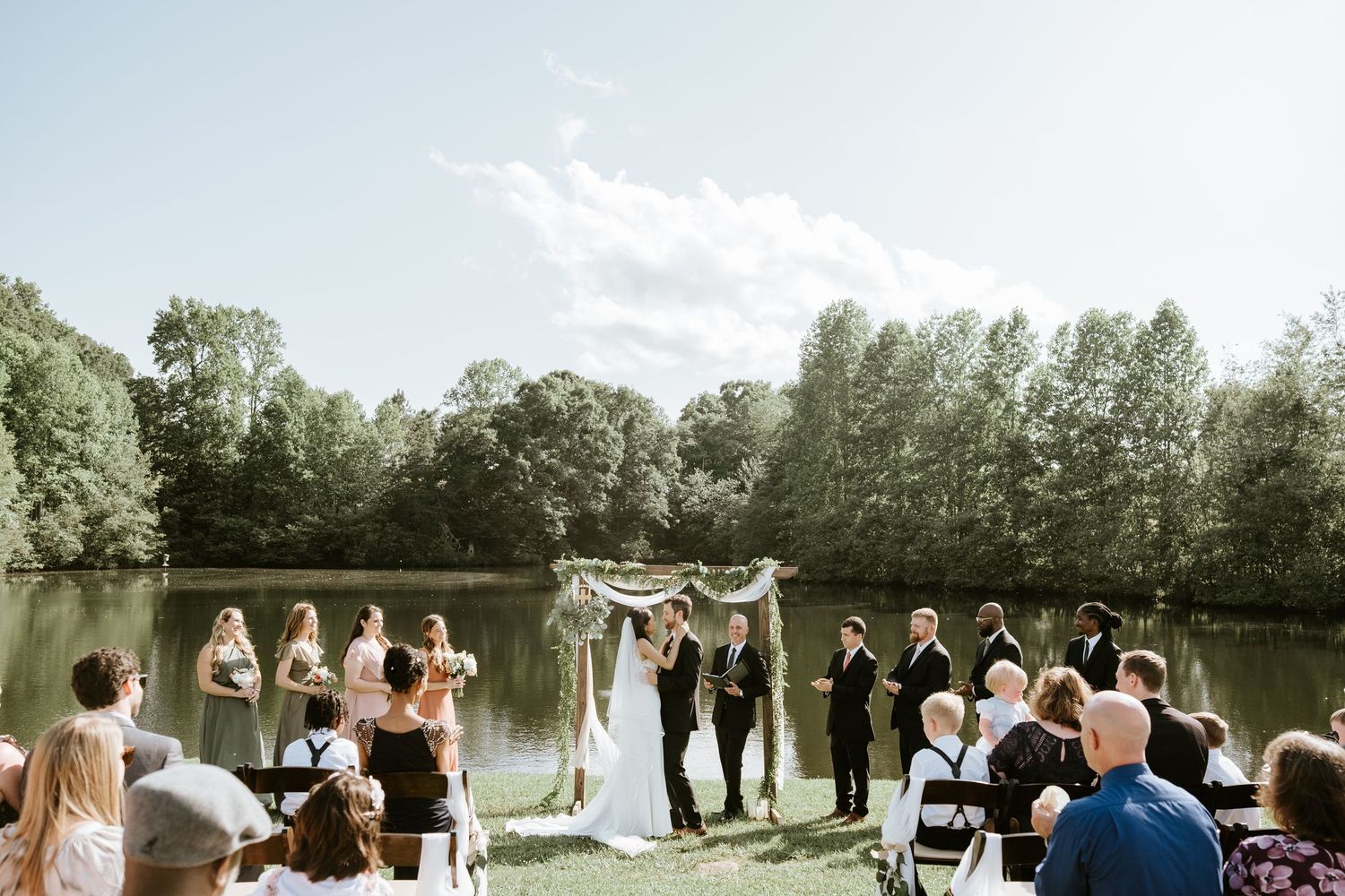 A wedding ceremony takes place outdoors by a serene lake with guests seated in white chairs and trees in the background.