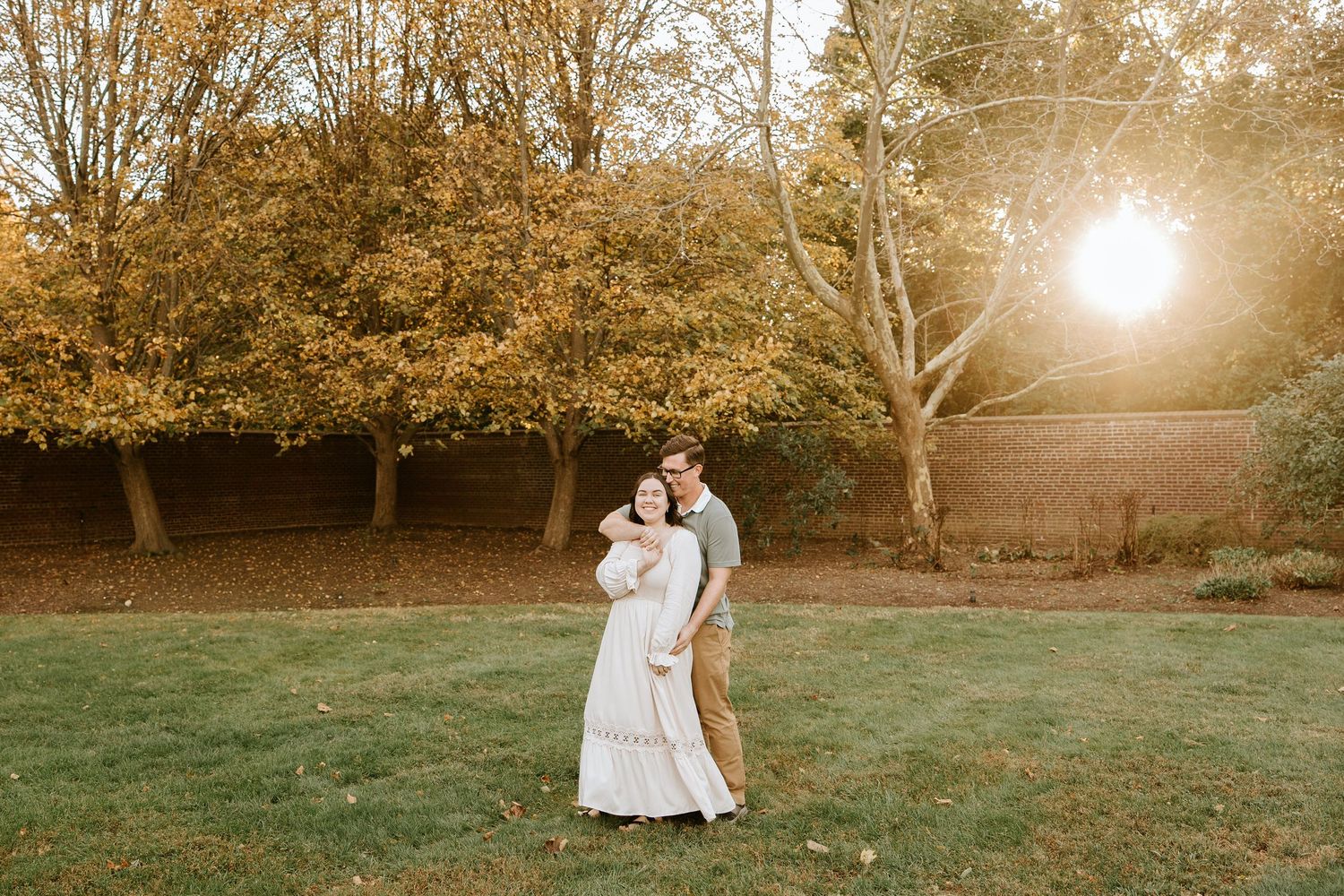 A couple in white outfits embraces in a sunlit meadow during golden hour as sunlight filters through autumn trees.
