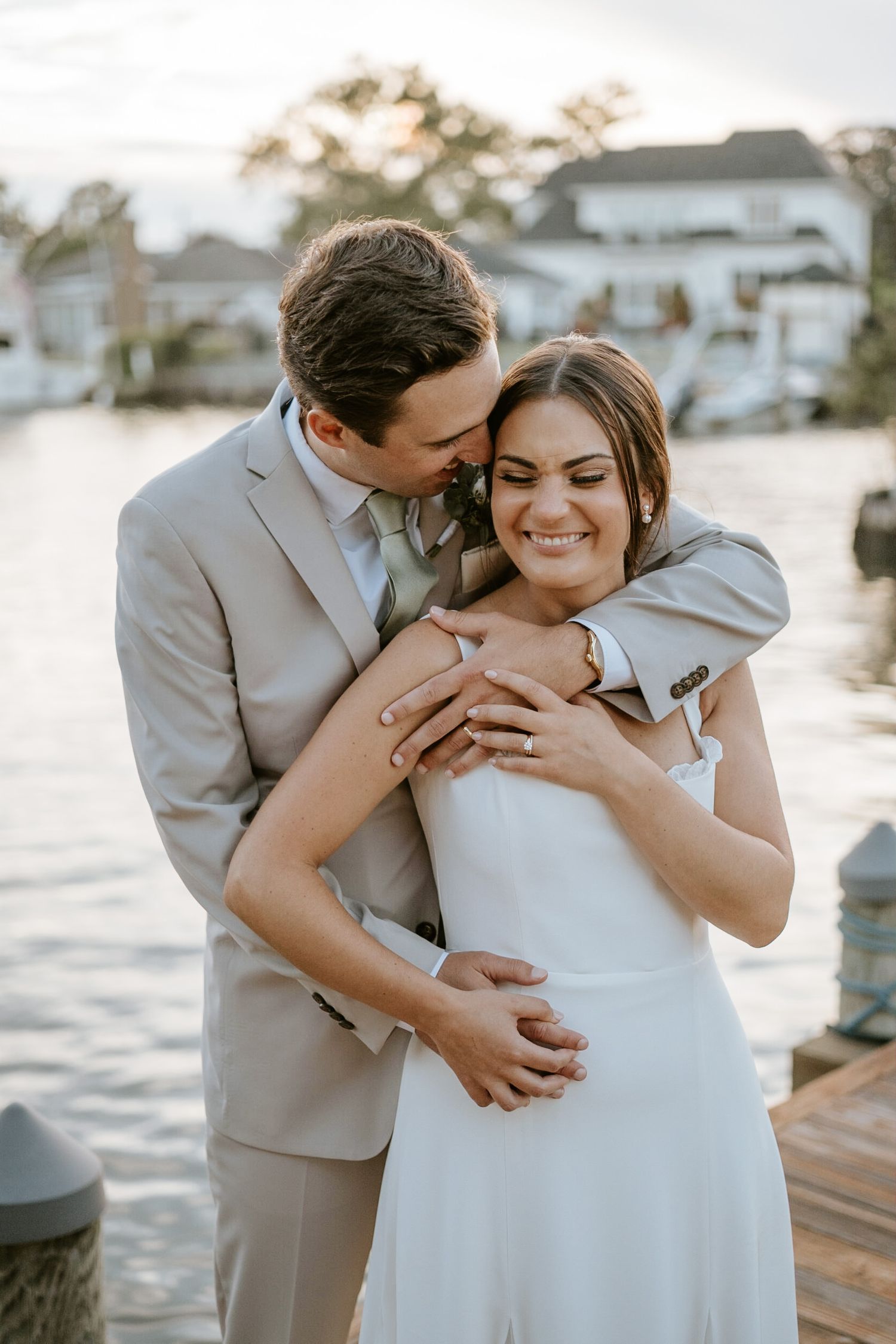 A groom in a gray suit embraces his smiling bride in a white dress on a waterfront dock at sunset.