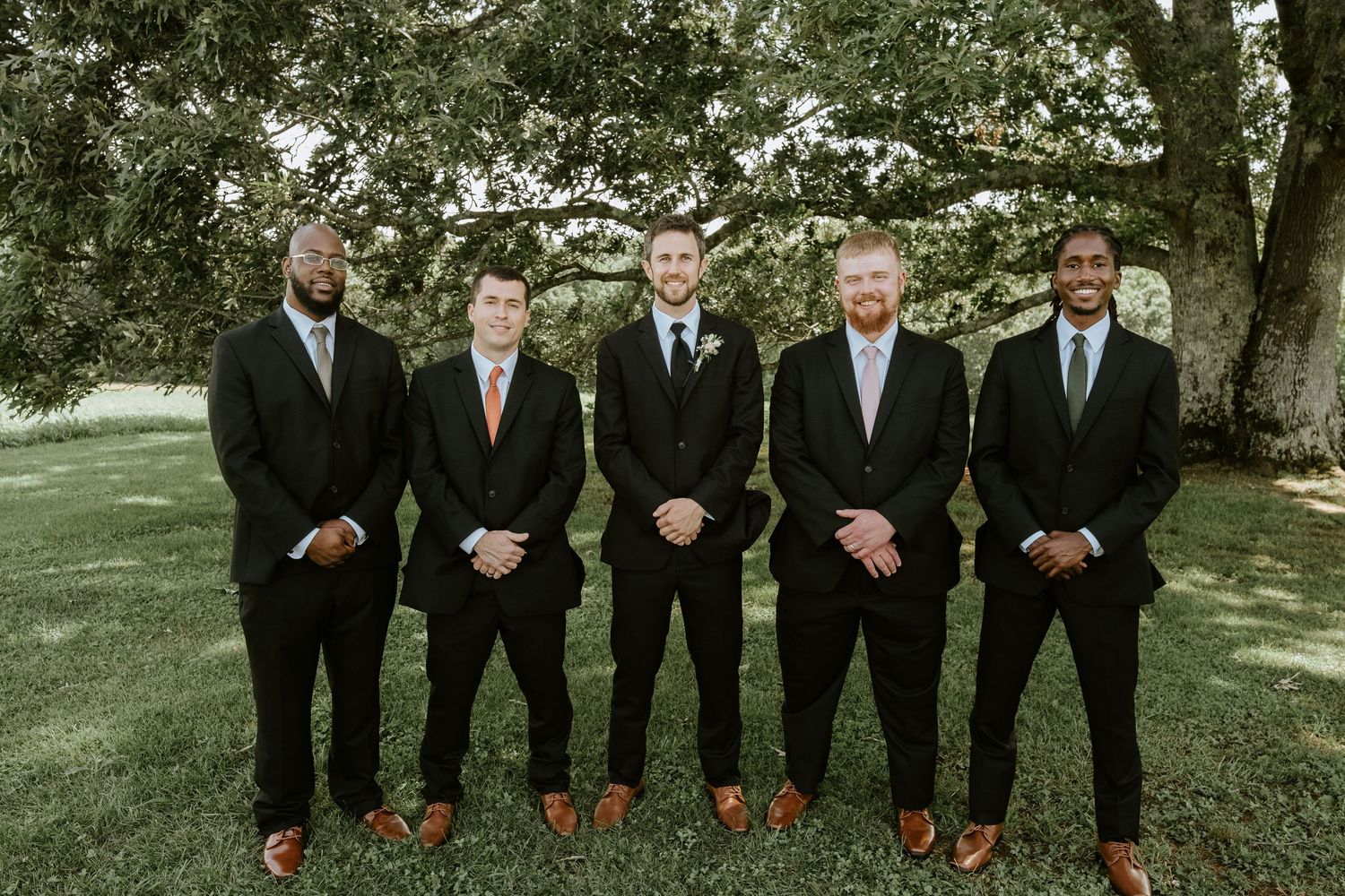 Groomsmen in black suits and red ties stand together in a line outdoors for a wedding photo.