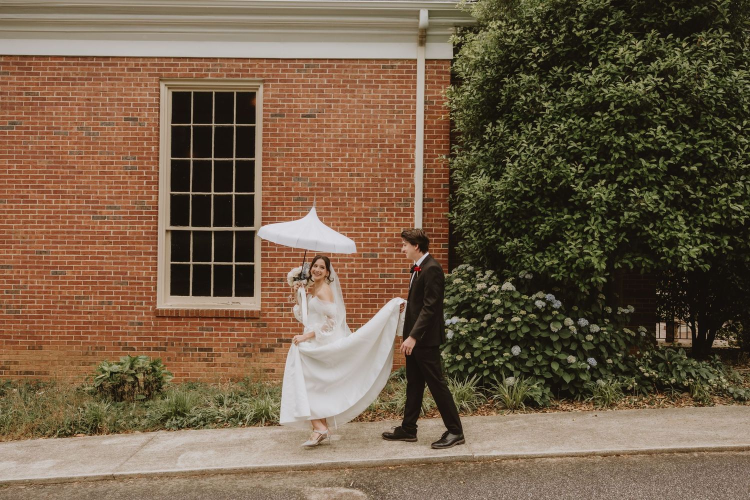 Bride and groom walk together outside a classic red brick building with white trim windows.