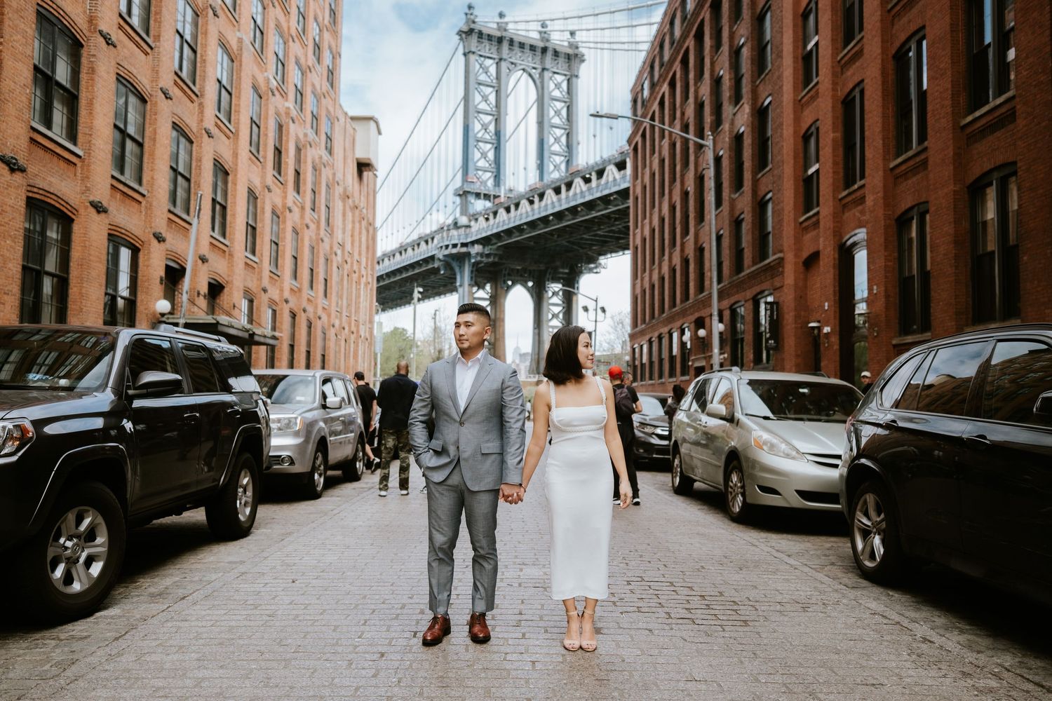 Urban street scene in DUMBO Brooklyn with Manhattan Bridge visible in background and parked cars along cobblestone road.