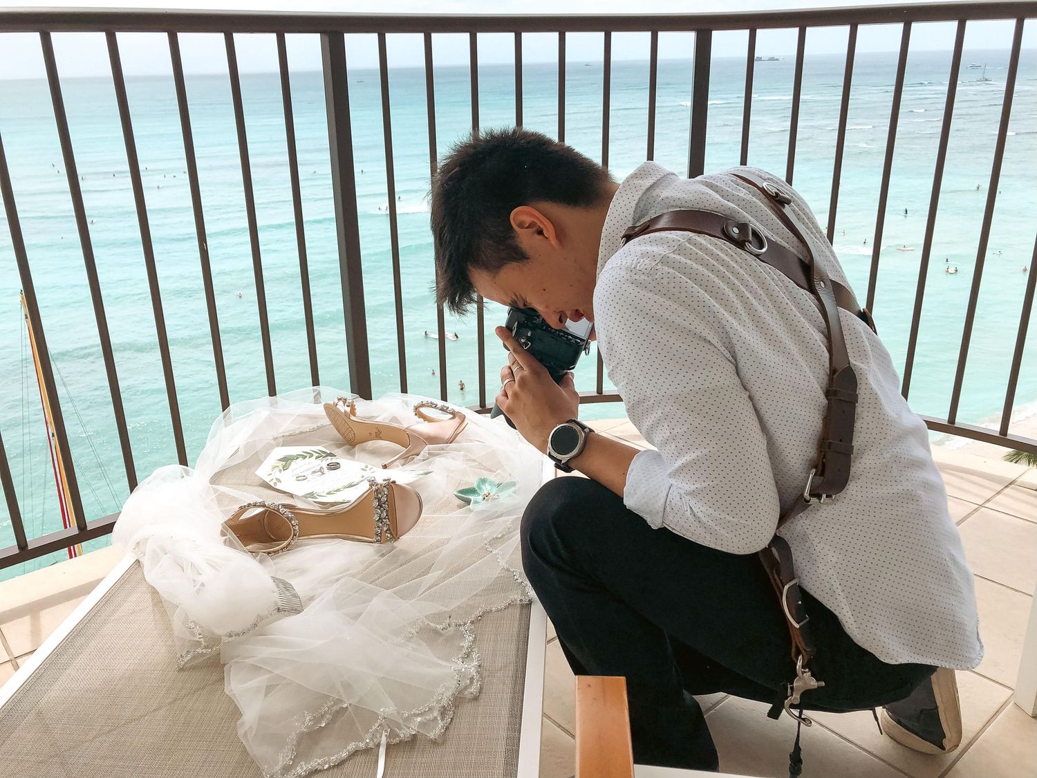 A man in a white shirt photographs wedding accessories on a balcony overlooking a turquoise ocean.