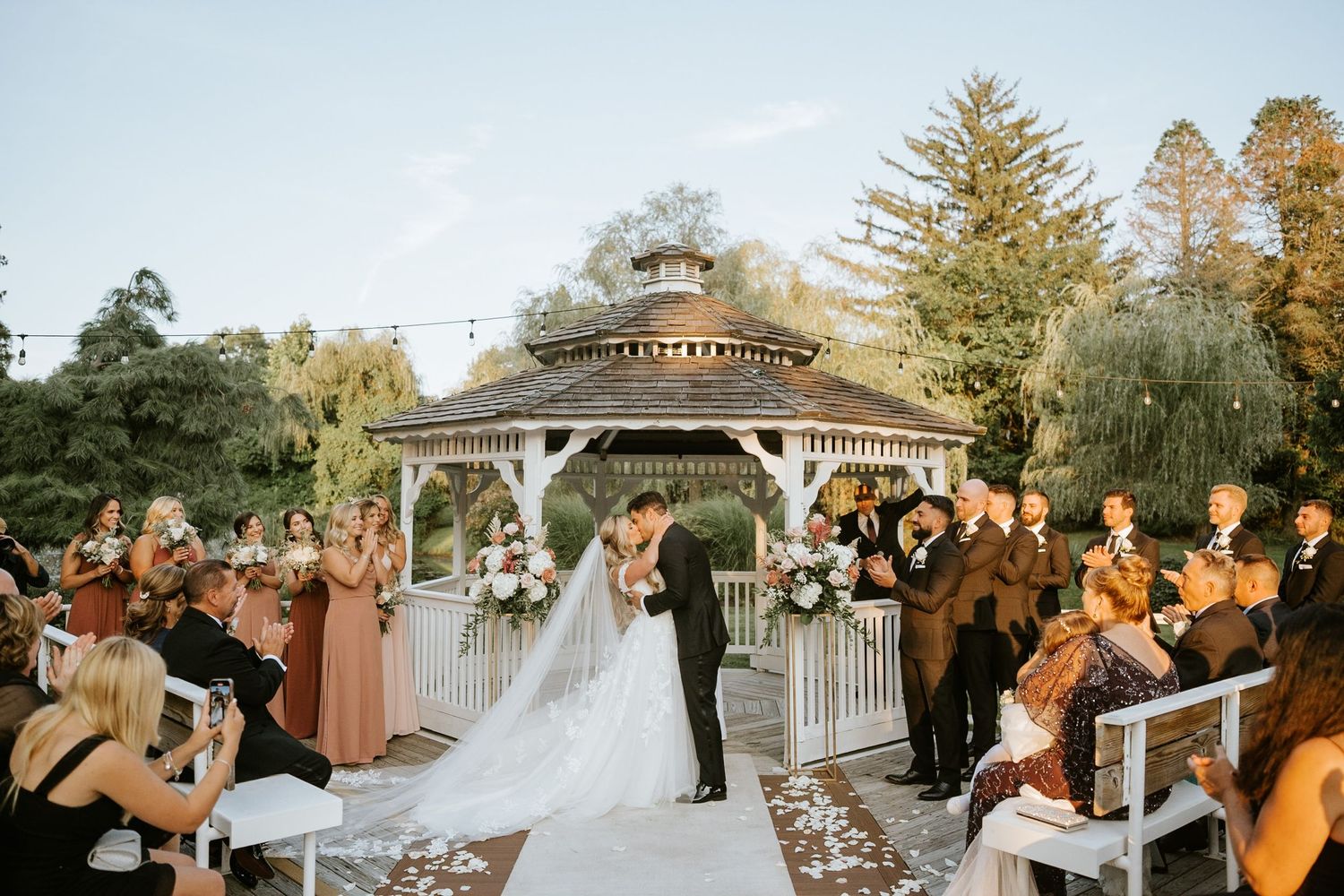 Couple shares first kiss at outdoor gazebo wedding ceremony with guests applauding on a sunny day.