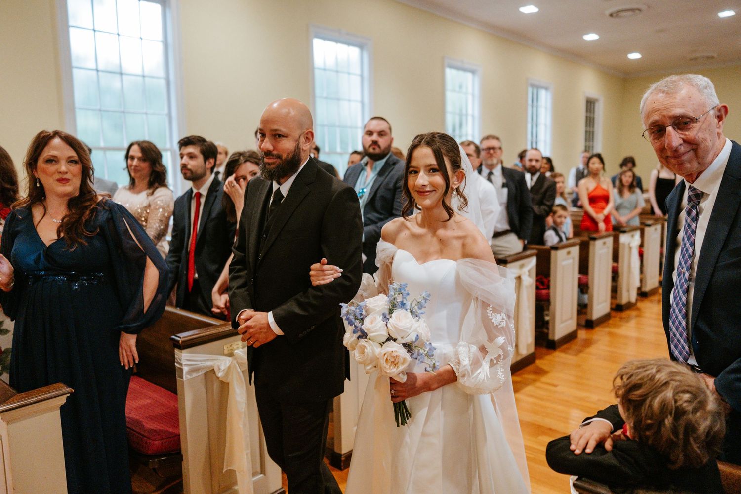 Wedding guests line the walls of a bright indoor venue as they watch an emotional ceremony unfold.