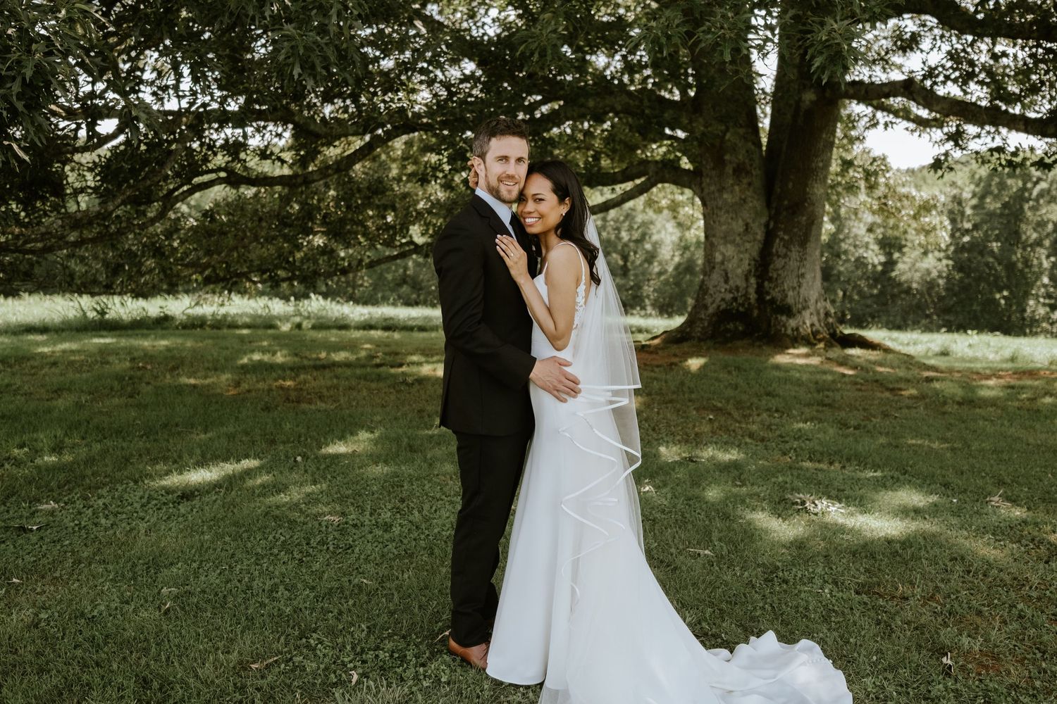 Bride and groom embracing outdoors under a large oak tree on lush green grass in wedding attire.
