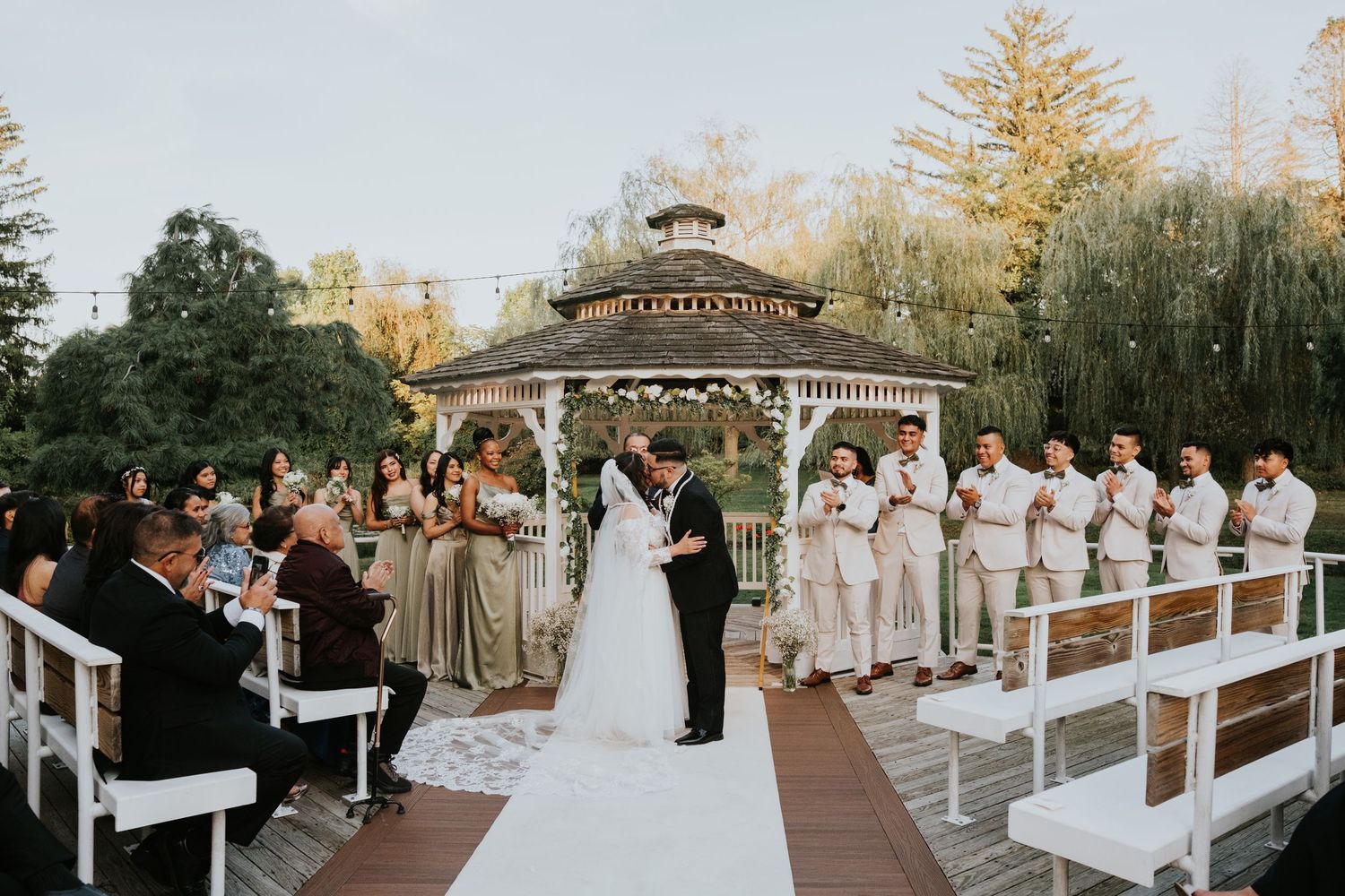 Outdoor wedding ceremony under a gazebo with bride and groom, bridesmaids in sage green, and groomsmen in beige suits.
