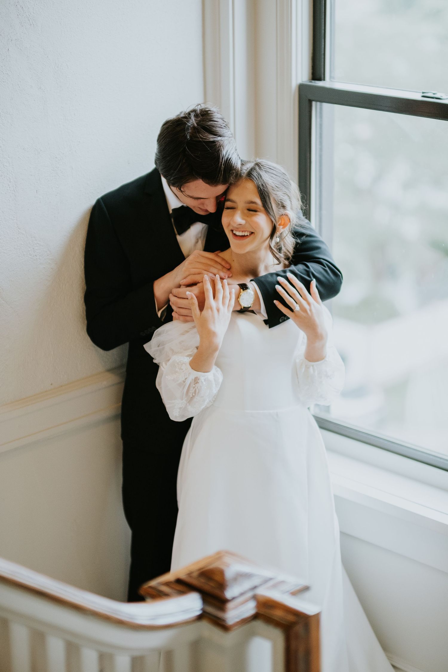 A romantic couple in formal attire share a tender moment by a bright window in an intimate indoor setting.
