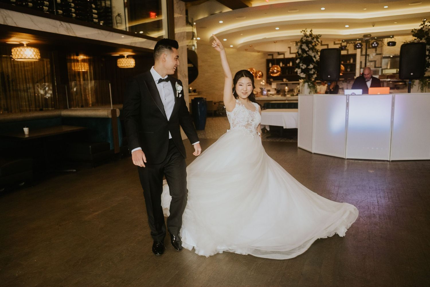 Bride in flowing white gown and groom in black tuxedo share first dance in elegant wedding venue.