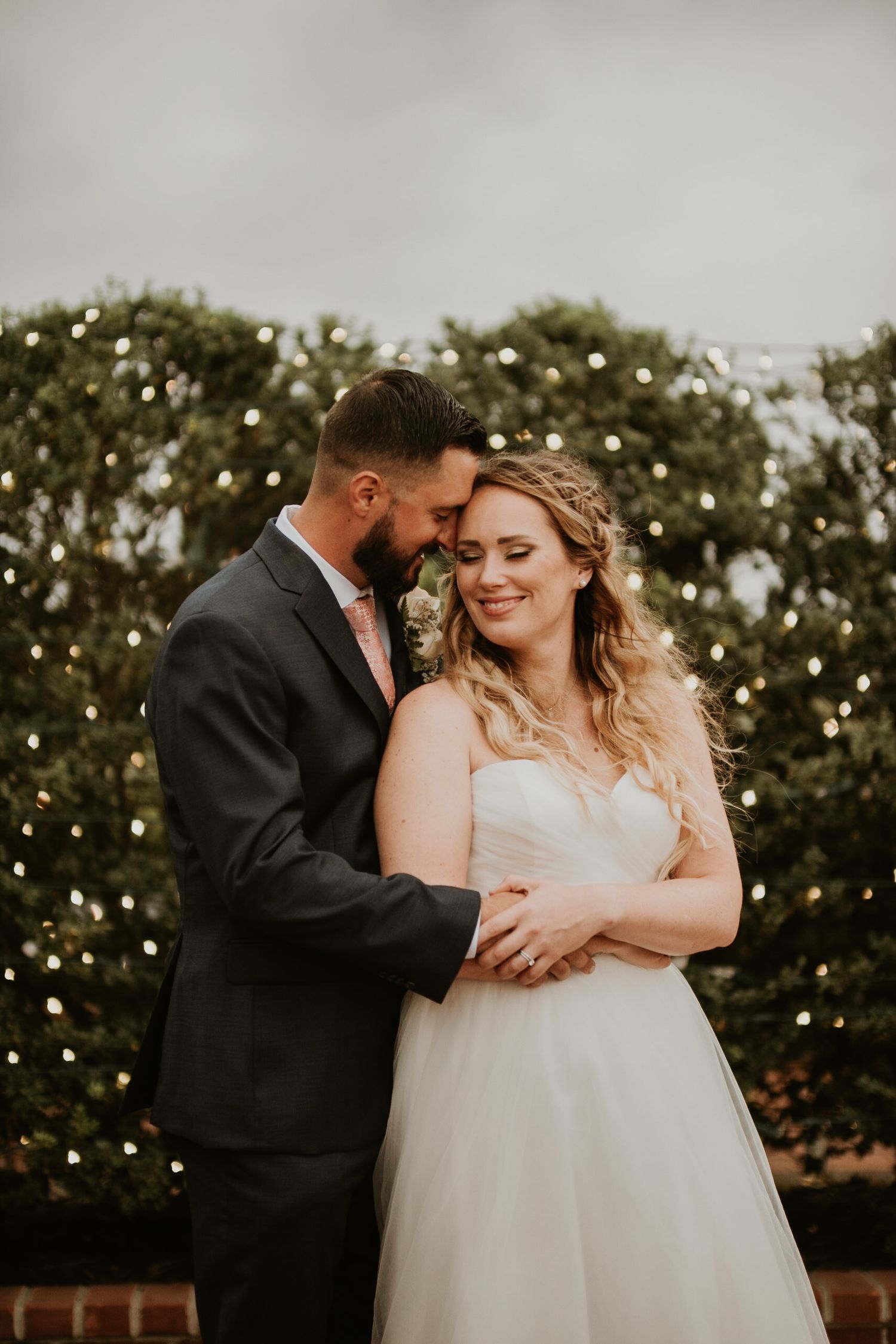 A bride in a white gown smiles as her groom in a gray suit embraces her in front of a hedge with twinkling lights.