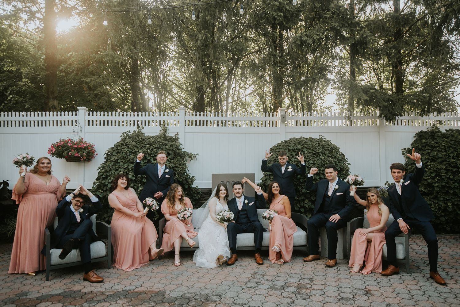 Wedding party in blush pink and navy attire posing on a garden bench with lush greenery and white fence backdrop.