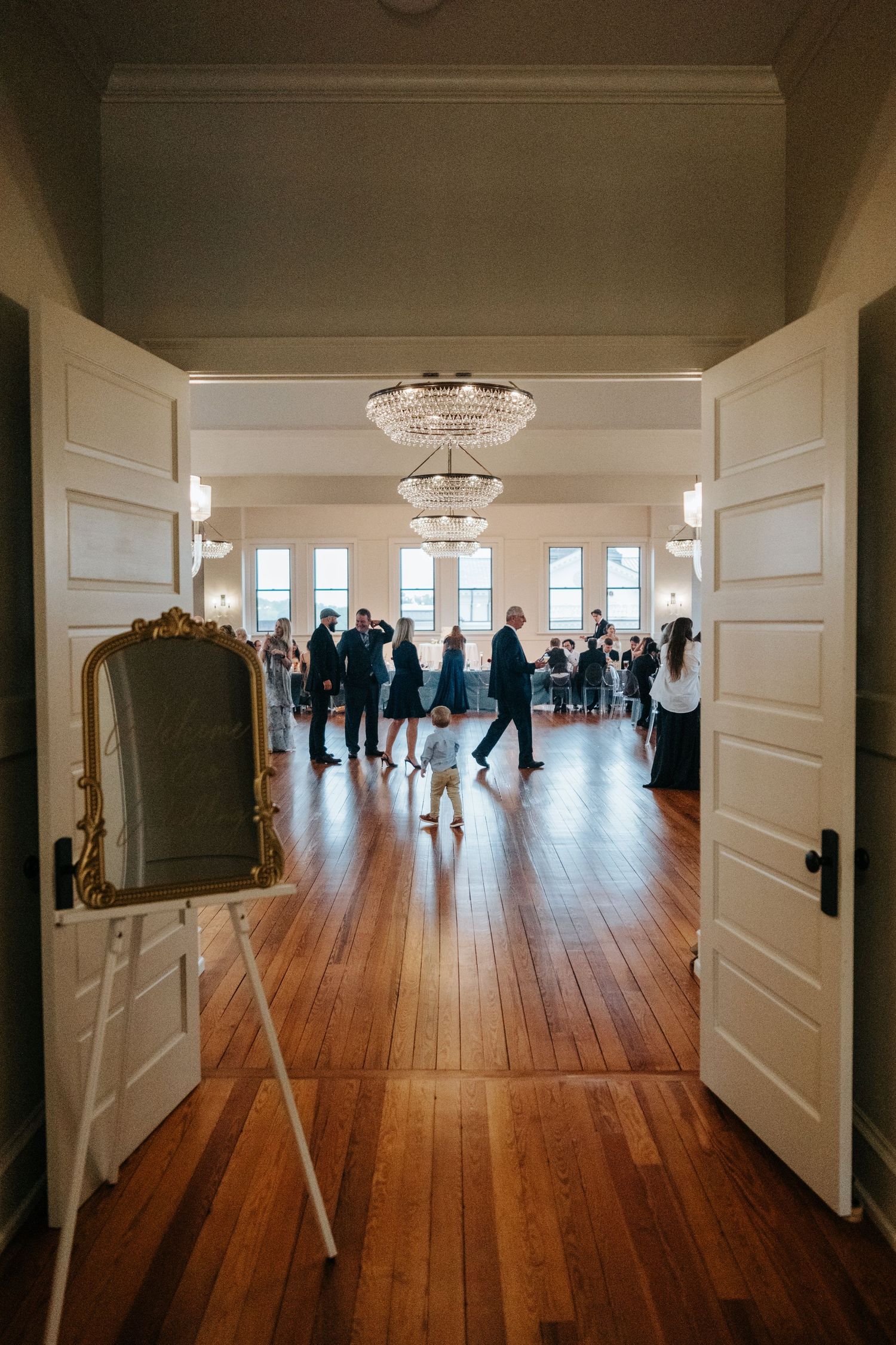 A long elegant hallway with white doors and wooden floors leads to a bright room where people gather in the distance.