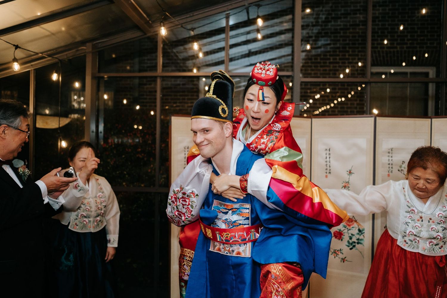 Couple in vibrant Korean hanbok attire celebrating at a wedding reception with guests in the background.