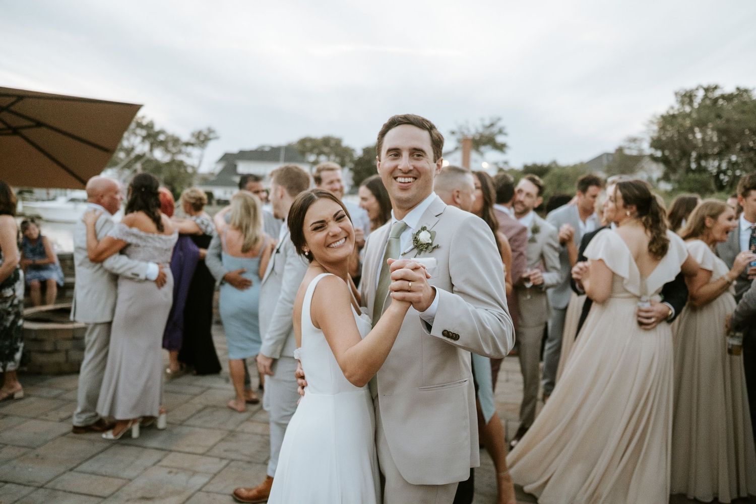 Bride and groom share first dance at outdoor wedding reception with guests dancing in background.