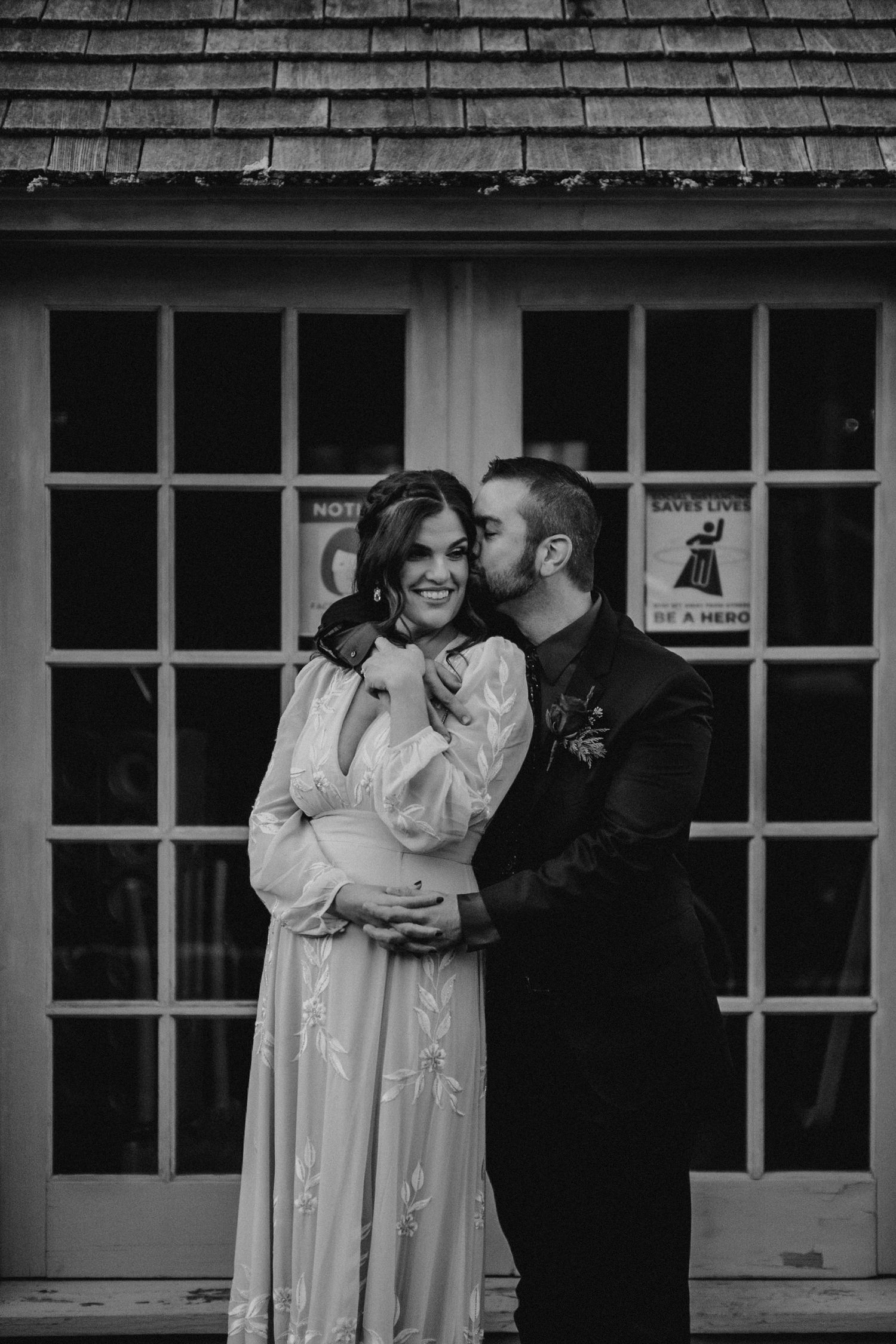Black and white photo of a couple, man kissing pregnant woman's cheek, both smiling, standing before a building with French doors.