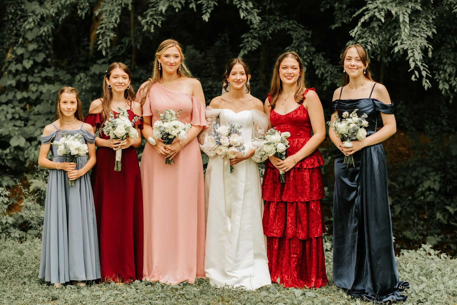 A bridal party in burgundy, pink, and gray dresses poses together outdoors holding bouquets.