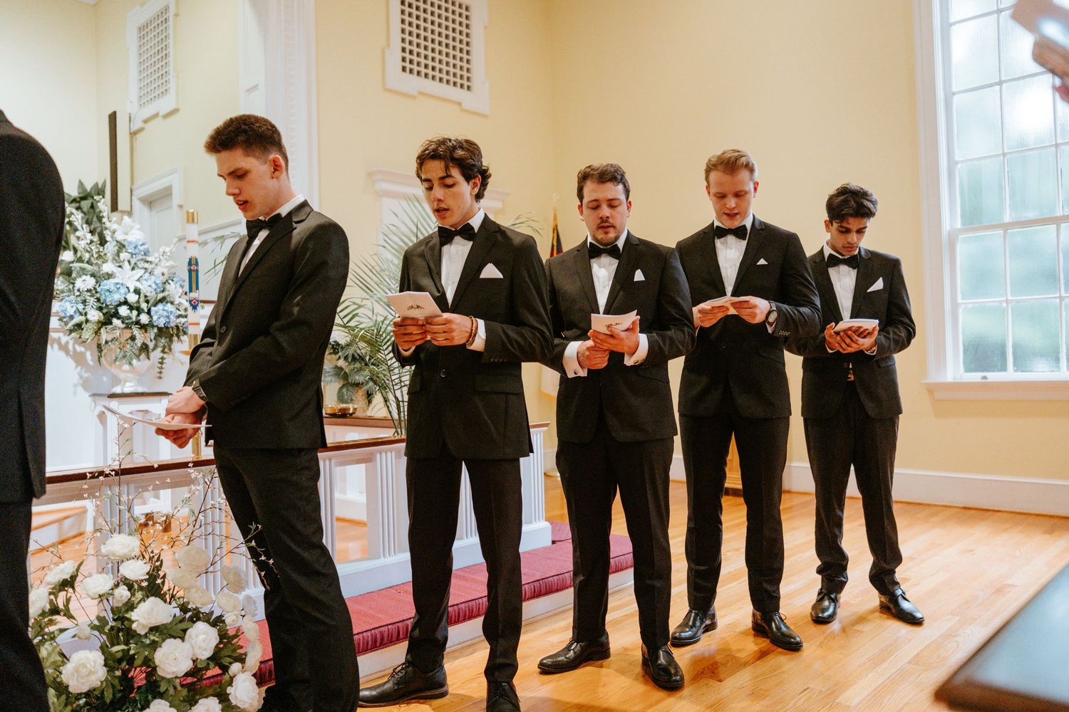 A row of groomsmen in black tuxedos check their watches in unison during a wedding photo.