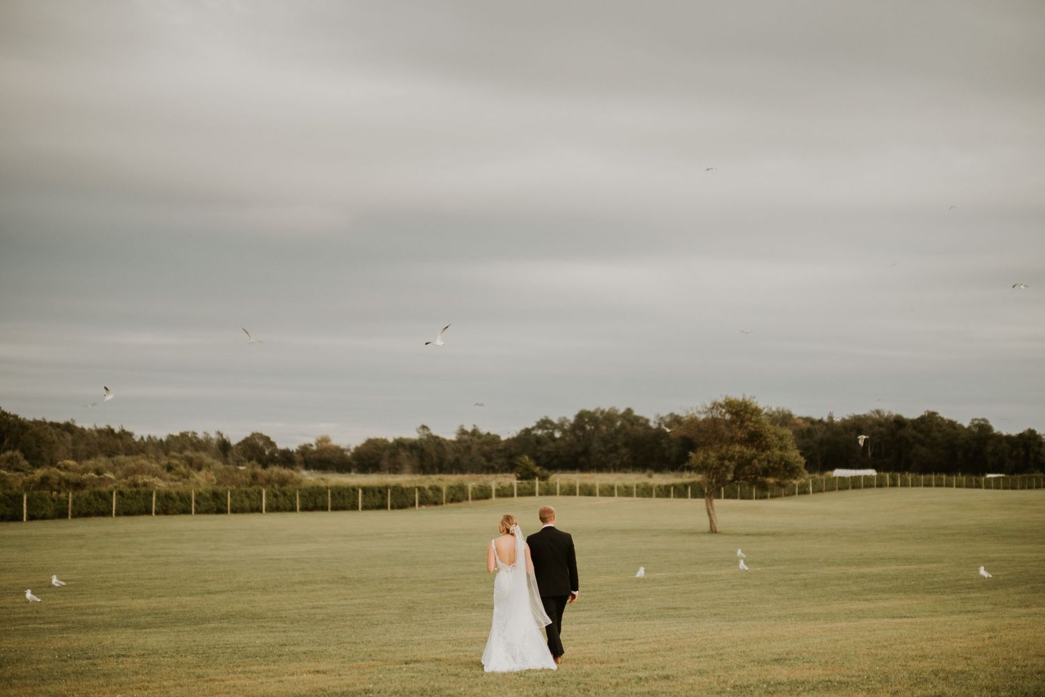 Bride and groom walking hand in hand across a lush green field on their wedding day under overcast skies.