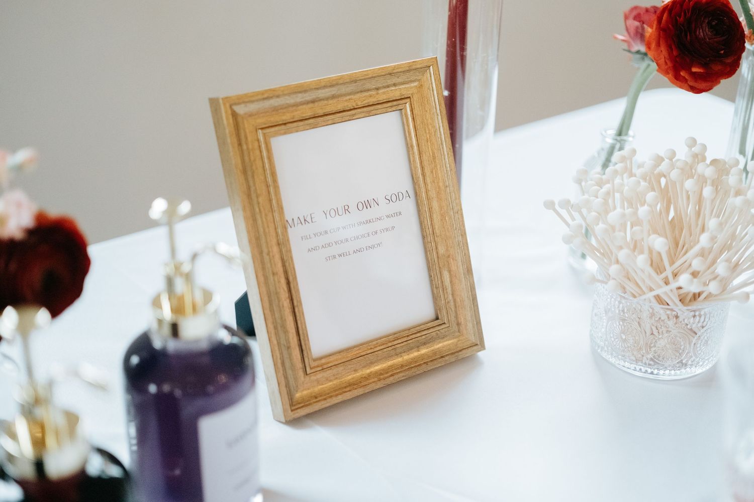 A wedding reception table setting features elegant white flowers, crystal glasses, and golden frames in a bright room.