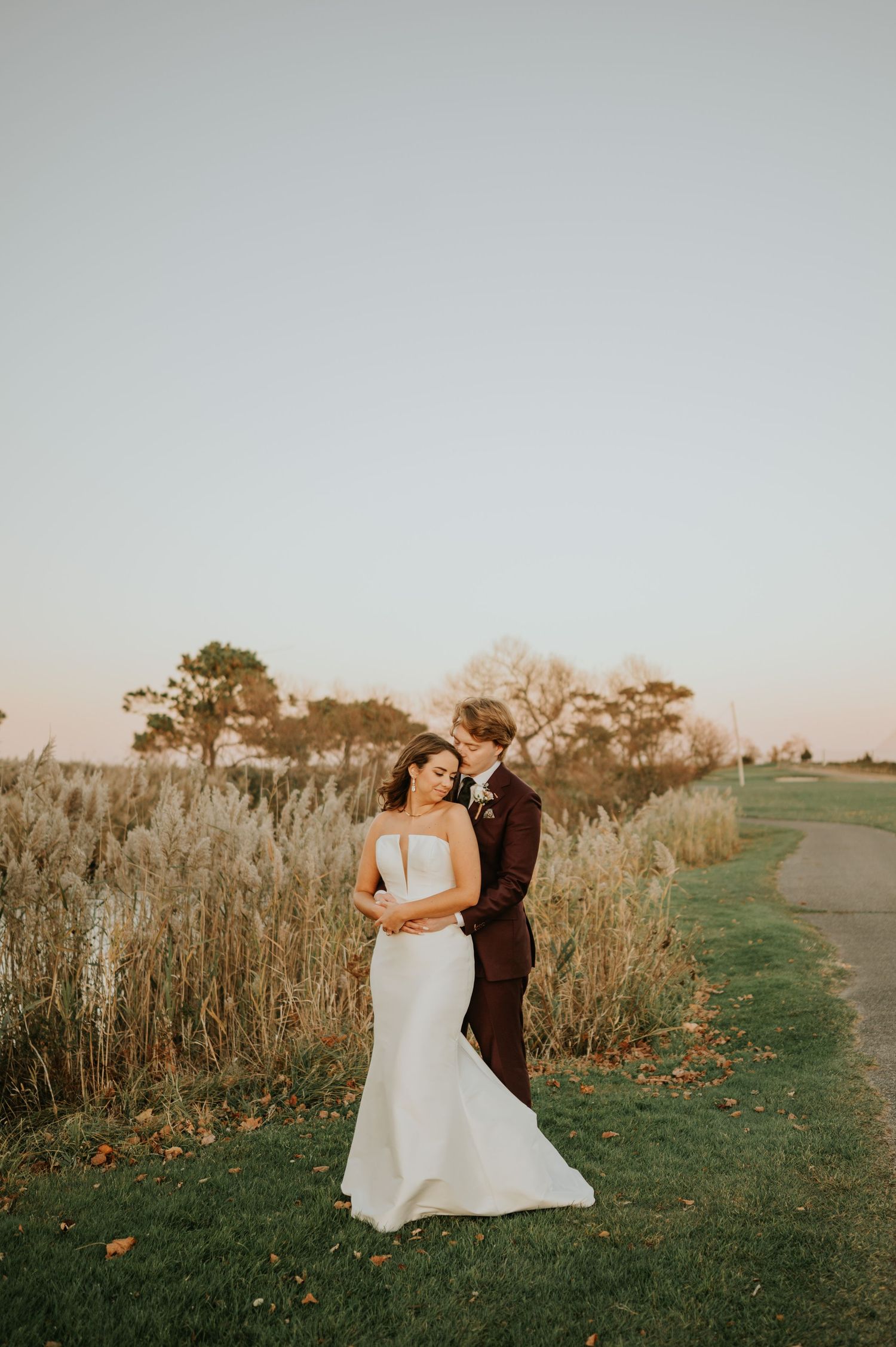 Bride and groom embrace at golden hour near tall grass and a pond on their wedding day.