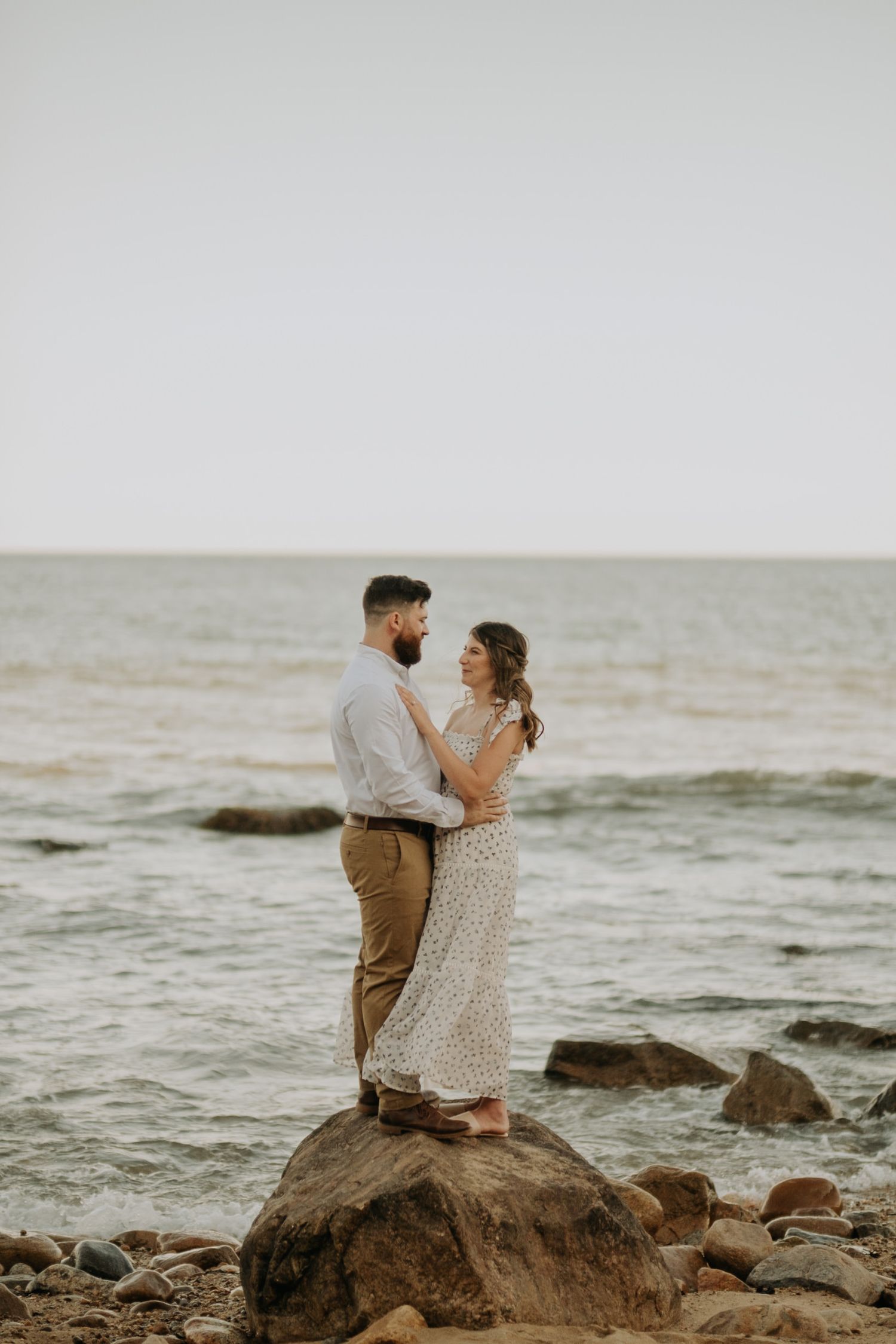 Couple embracing on rocky beach shore, standing on a large rock with ocean waves in the background.
