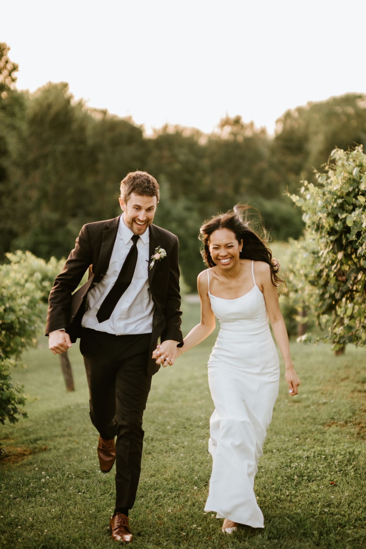 Happy newlyweds hold hands running through a vineyard, bride in white gown and groom in black suit on their wedding day.