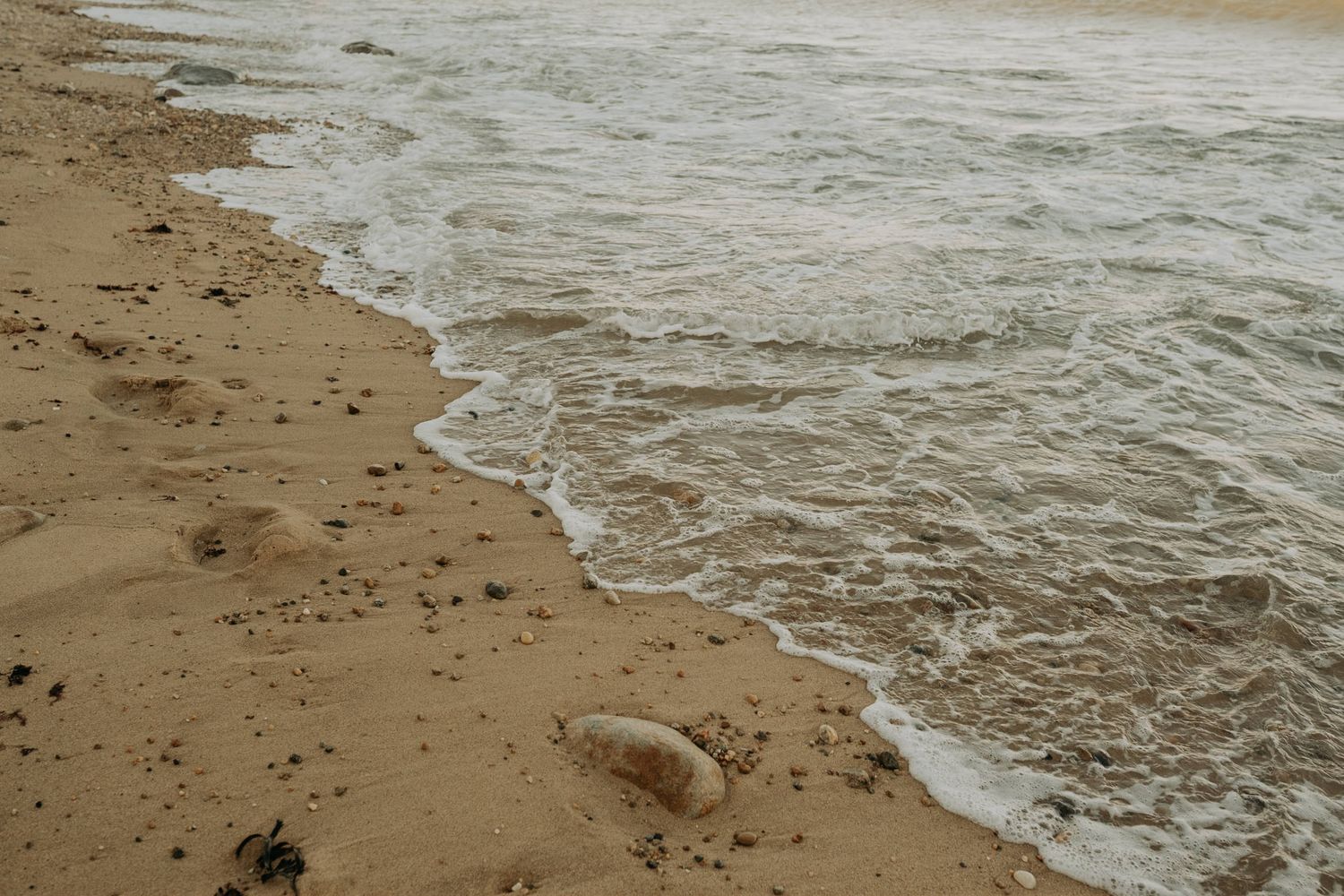 Gentle ocean waves washing onto a sandy beach shore with scattered pebbles and footprints in wet sand.