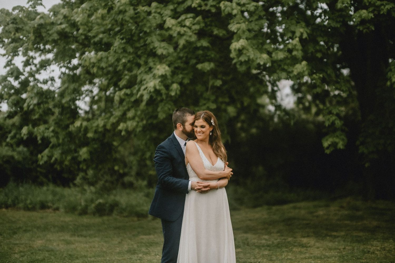 Couple embracing outdoors on green lawn with lush trees, bride in white gown and groom in navy suit at garden wedding.