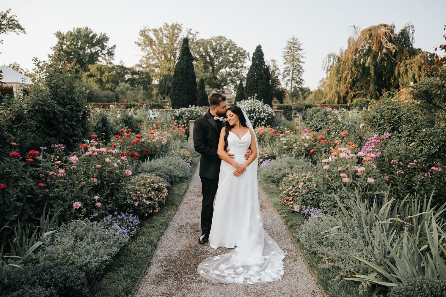 Bride and groom embrace on a garden path surrounded by colorful roses and lush greenery at an outdoor wedding.